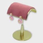Drop pearl earrings with pink and gold accents displayed on a modern gold stand for MEENA product page.