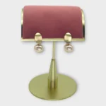 A pair of Auralis Drop earrings with gold oval tops and pearl drops displayed on a pink and gold stand.