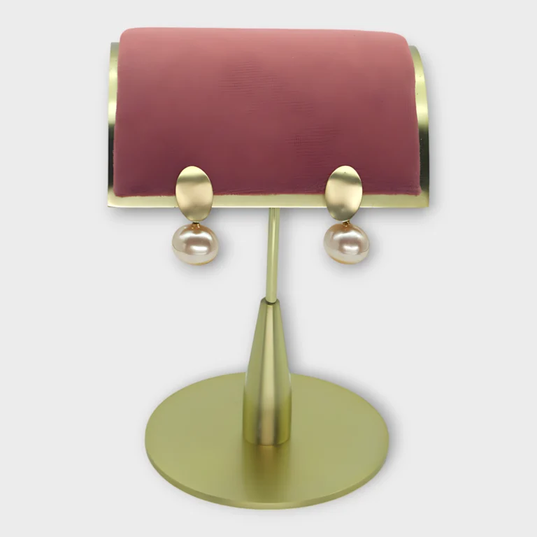 A pair of Auralis Drop earrings with gold oval tops and pearl drops displayed on a pink and gold stand.