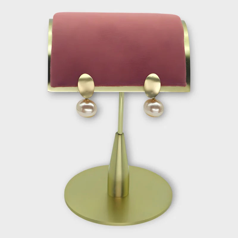 A pair of Auralis Drop earrings with gold oval tops and pearl drops displayed on a pink and gold stand.