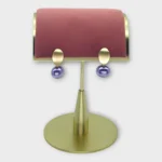 A pair of Auralis Drop earrings with gold oval tops and purple dangling beads on a gold display stand.