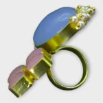 Angle view of STONIRA adjustable statement ring featuring a large Blue Chalcedony gemstone, Rose Quartz accents, pearl detailing, and a gold-tone setting.