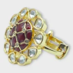 Angle view of STONIRA gold cocktail ring featuring a central glossy crimson red domed stone with a gold grid overlay and an outer halo of clear teardrop gems