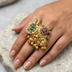 Women wearing STONIRA adjustable gold temple charm ring featuring red gemstone, pearl accents, intricate heritage motifs and ghungroo detailing for festive and wedding wear.