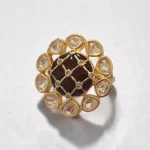 STONIRA gold cocktail ring featuring a central glossy crimson red domed stone with a gold grid overlay and an outer halo of clear teardrop gems