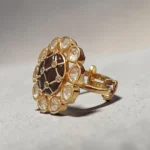 Side view of STONIRA gold cocktail ring featuring a central glossy crimson red domed stone with a gold grid overlay and an outer halo of clear teardrop gems