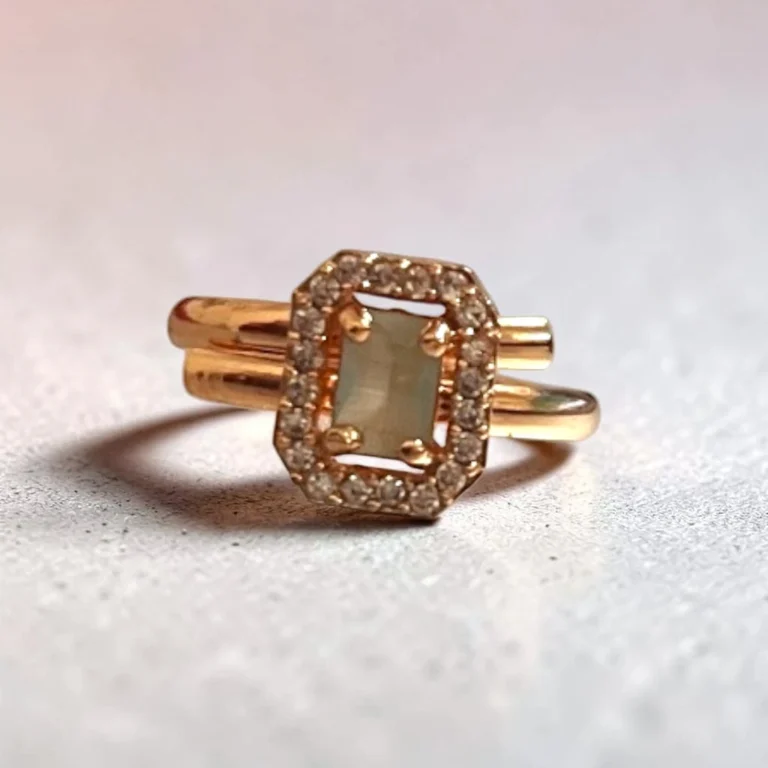 Angle-view-of-STONIRA-rose-polish-statement-ring-featuring-a-rectangular-sage-green-gemstone-enclosed-in-an-octagonal-halo-of-shimmering-clear-crystals-on-a-neutral-backdrop.