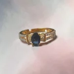 Elegant STONIRA gold-toned adjustable cocktail ring displaying a central oval navy blue sapphire-colored gemstone flanked by baguette crystals on a white backdrop.