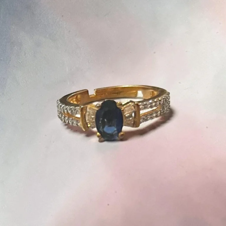 Elegant STONIRA gold-toned adjustable cocktail ring displaying a central oval navy blue sapphire-colored gemstone flanked by baguette crystals on a white backdrop.