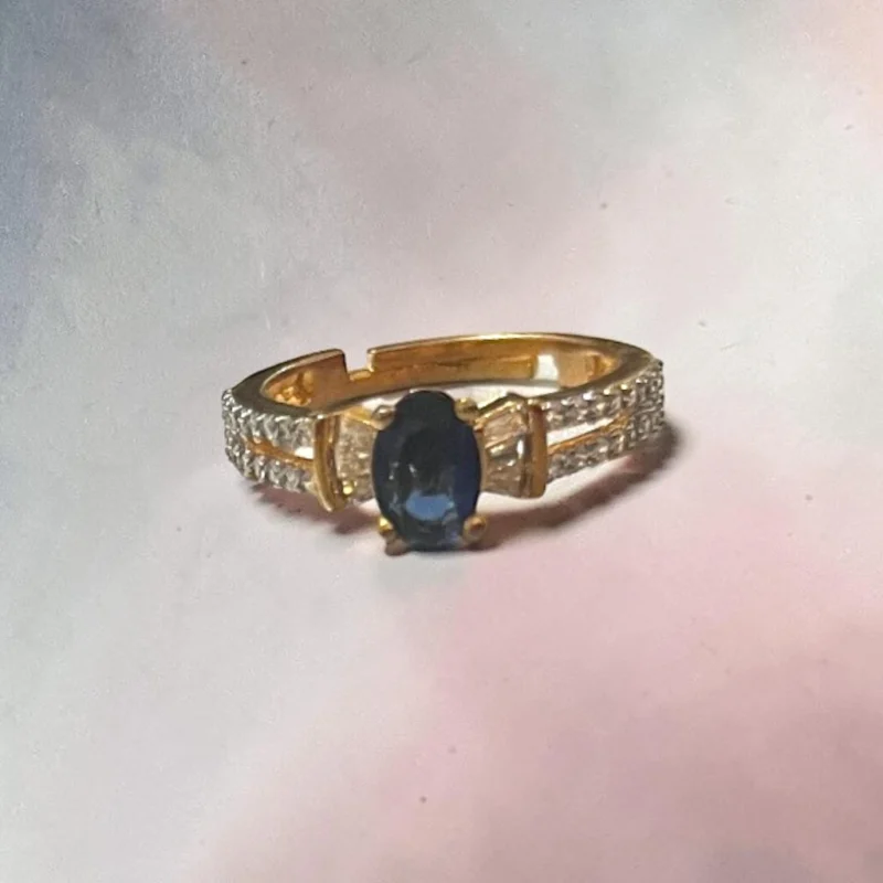 Elegant STONIRA gold-toned adjustable cocktail ring displaying a central oval navy blue sapphire-colored gemstone flanked by baguette crystals on a white backdrop.