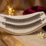 STONIRA-handcrafted-broad-oxidized-silver-payal-set-with-ruby-red-stones-and-dense-ghungroo-bells-displayed-on-a-velvet-surface
