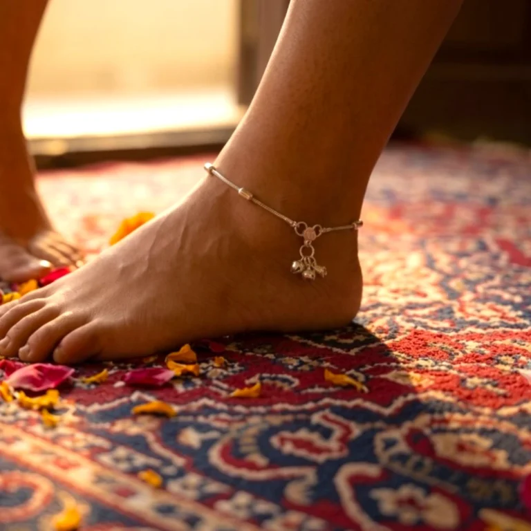 Side-profile-close-up-of-a-womans-foot-walking-on-a-rich-patterned-red-rug-scattered-with-marigold-petals-wearing-the-STONIRA-silver-bead-payal-anklet-with-hanging-musical-bells.